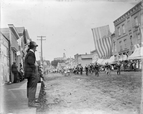 View from wood sidewalk towards a marching band parading up Main Street during a patriotic celebration. There is a banner that reads: "Welcome" in the background. The American Flag hanging over the street in the foreground has 45 stars.