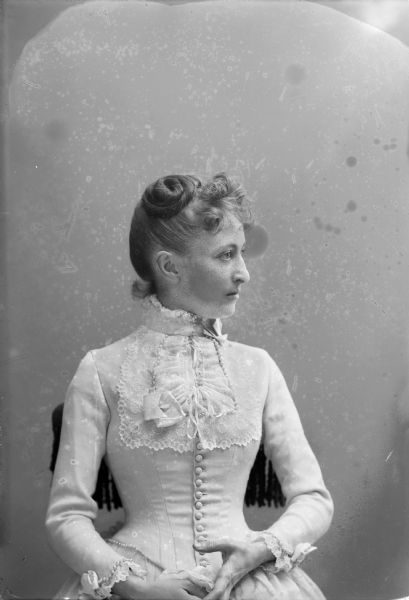 Waist-up studio portrait of Lottie Porter, wife of Frank Long, sitting in a chair. Her head is turned to the right in profile. She wears a high-collared white dress with lace and pearls at the neckline and sleeves.