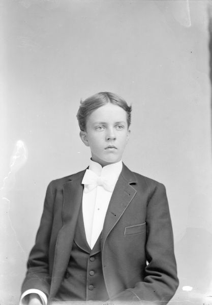 Waist-up studio portrait of young European American man posed sitting and wearing a dark-colored suit coat, vest, and light-colored bow tie. Identified as Price Powers.