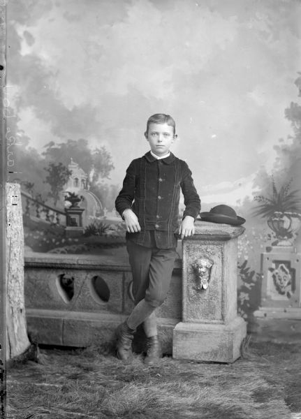 Full-length studio portrait in front of a painted backdrop of a European American boy posed standing with his left hand resting on a prop stone wall and column. A carpet of grass is under his feet. There is a hat on top of the column, and he wears a dark-colored button-down jacket and knickers. Boy identified as probably the son of L.H. Clark.