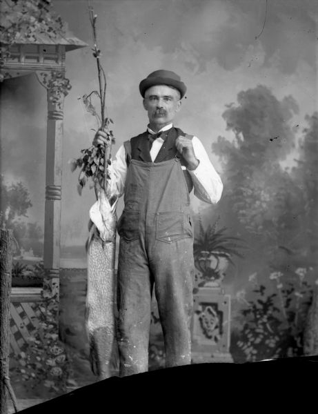 Studio portrait in front of a painted backdrop of European American man posed standing. He is holding up in his right hand a fishing pole, tree branch, and muskellunge. He is wearing overalls over a dark-colored vest, shirt, bow tie, and hat. Man identified as probably John Wiggin.