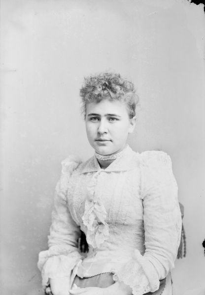 Waist-up studio portrait of European American woman posed sitting and wearing a light-colored dress with a lace and ruffle bodice and a choker necklace. Woman identified as Mary O. Mills.