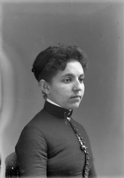 Waist-up studio portrait of young European American woman posed sitting facing to the right. She is wearing a dark-colored, close fitting button-down coat, with a collar pin and hanging brooch. Woman identified as Minnie Taylor.