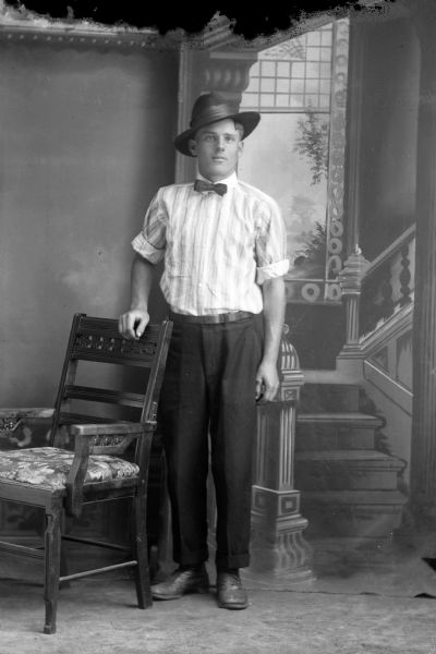 Full-length studio portrait in front of a painted backdrop of a European American man posed standing with his right hand on the back of a wooden chair. He wears a light-colored striped shirt, dark-colored trousers, bow tie, and hat. Man identified as probably John Darwin.