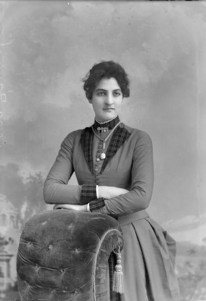Three-quarter length studio portrait in front of a painted backdrop of a European American woman posed standing partially behind a chair. She is resting her arms on the back of the upholstered chair, and she wears a dark-colored dress with a button-down bodice, darker-colored plaid trim, chain ribbon necklace with a locket, and a collar pin. The woman is identified as probably Lena Baumel.