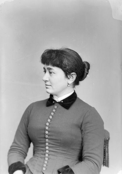 Waist-up studio portrait of a European American woman with a hair bun posed sitting and looking towards the left. She is wearing a light-colored dress with dark-colored trim and a button-down bodice, collar pin, and earrings. The woman is identified as Mary Wright, from the class of 1882.