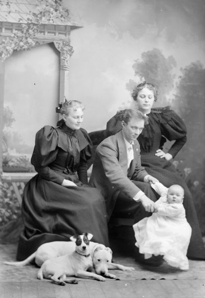 Studio portrait in front of a painted backdrop of a European American family and their two dogs. One woman is posed sitting on the left with her hands in her lap, looking at the baby that a man, center, is balancing on his foot. There is a woman sitting on the chair arm on the right and she is looking down at the infant. Two dogs are laying on the floor in front of the group. Group is identified, from left to right, as probably the wife of Dr. Eugene Krohn, Eugene Krohn, Victor Krohn, and the sister of Mrs. Krohn.