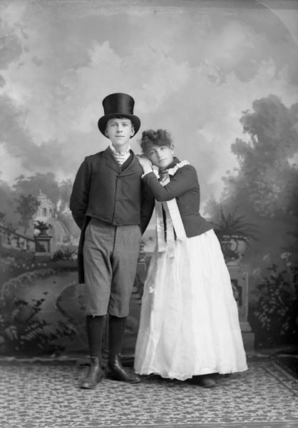 Full-length studio portrait in front of a painted backdrop of a young European American man and woman posed standing. The woman,&#65279;&#65279; wearing a light-colored skirt, dark-colored coat, and light-colored ribbons is leaning on the left shoulder of the man. The man is wearing a suit coat with tails, knickers, striped necktie and a top hat. Identified as probably Will Catchpole on the left, and Florence Wickland on the right.