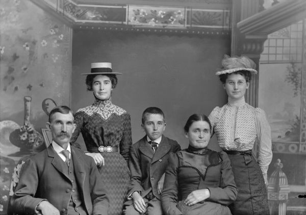 Studio portrait in front of a painted backdrop of a European American family. On the left a man, John Teadman, Sr., is sitting and wearing a dark-colored suit coat, vest, and necktie. A woman, Nellie Teadman Roberts, wife of J. Roberts, is standing behind him. She is wearing a dark-colored zig-zag pattern dress with light-colored collar and a light-colored straw hat. In the center is a boy, John Teadman, Jr., sitting and wearing a dark-colored double-breasted suit coat and neckerchief. Next to the boy is a woman, the wife of John Teadman, sitting and wearing a dark-colored dress and collar pin. On the far right a woman, Laura Teadman Woodford, wife of R. Woodford, is standing and wearing a dark-colored skirt, light-colored striped blouse, and light-colored hat.