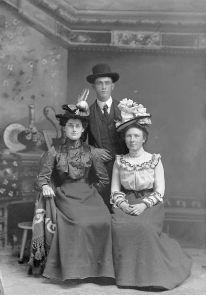 Studio portrait in front of a painted backdrop of two European American women and a European American man. The woman sitting on the left is wearing a dark-colored dress with a button-down bodice, eyeglasses, and a hat with feathers. The man standing in the center is wearing a dark-colored suit coat, vest, necktie, tie pin, and hat. The woman sitting on the right is wearing a dark-colored skirt, light-colored blouse with ruffled trim, and a hat. Identified from left to right as probably the wife of Robert Woodford, Robert Woodford, and Nina Woodford.
