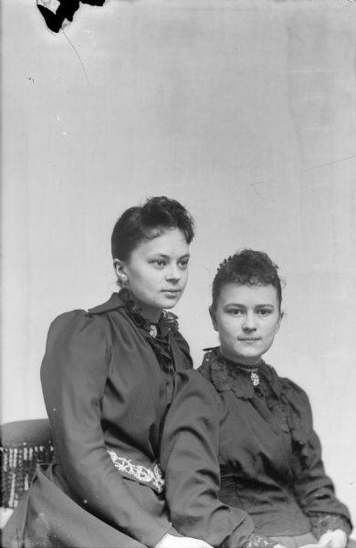 Studio portrait of two European American women sitting on a chair. The woman on the left is sitting on the arm of the chair and is wearing a dark-colored dress with lace trim and a collar pin. The woman on the right is wearing a dark-colored dress with lace trim, collar pin, and choker necklace. Identified from left to right as probably Wally Hirsch and Stella Hirsch.