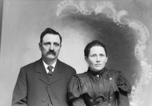 Waist-up studio portrait in front of a painted backdrop of a European American man with a moustache and a European American woman. The man is sitting on the left and is wearing a dark-colored suit coat and necktie. The woman is sitting on the right is wearing a dark-colored dress with billowy sleeves, collar pin, and brooch. Identified from left to right as probably L. Arnold and his wife.