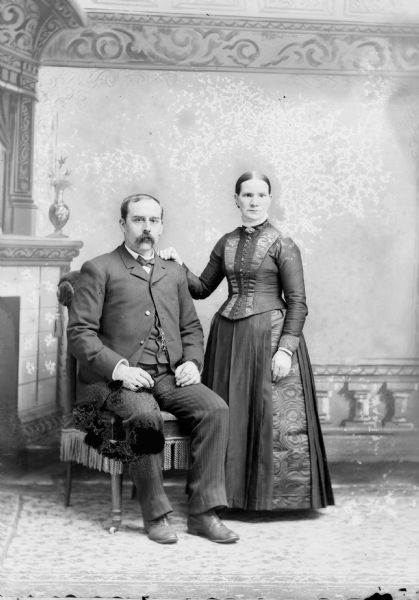 Studio portrait in front of a painted backdrop of a European American man and woman. The man has a moustache and is sitting in a chair on the left and is wearing a dark-colored suit coat, trousers, vest, and necktie. The woman is standing on the right with her right hand on the left shoulder of the man. She is wearing a dark-colored dress with a button-down bodice. Identified from left to right as Gilbert Melby and his wife.