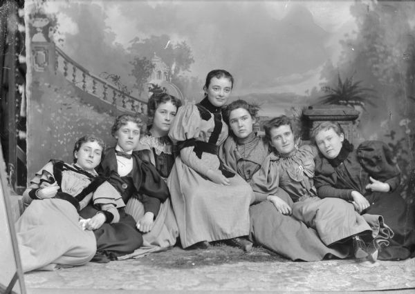 Studio portrait in front of a painted backdrop and prop column of seven European American women sitting on the floor. They are all leaning against one another. Identified from left to right as probably Grace Ogden, Jean Wright, Blanche Argyle, Ella Stiehl, Blanche Gund, Jean Wright Victor, and Grace Robie.