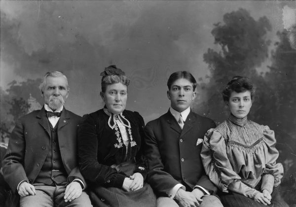 Three-quarter length studio portrait of a European American family posed sitting in front of a painted backdrop. A middle-aged man with a moustache and beard is on the far left wearing a dark-colored suit coat, vest, checked trousers, bow tie, and watch chain. Second from the left is a middle-aged woman wearing a dark-colored dress with light-colored embroidered trim. Next is a young man wearing a dark-colored suit coat and necktie. On the far right is a young woman wearing a ruffled blouse and a skirt. Identified from left to right as J.R. Ogden, Mrs. J.R. Ogden, Carl Ogden, and Grace Ogden.