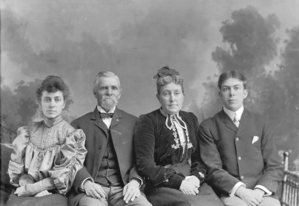 Three-quarter length studio portrait of a European American family posed sitting in front of a painted backdrop. On the far left is a young woman wearing a ruffled blouse and a skirt. Second from left is a middle-aged man with a moustache and beard wearing a dark-colored suit coat, vest, checked trousers, bow tie, and watch chain. Second from the right is a middle-aged woman wearing a dark-colored dress with light-colored embroidered trim. On the far right is a young man wearing a dark-colored suit coat and necktie. Identified from left to right as Grace Ogden, J.R. Ogden, Mrs. J.R. Ogden, and Carl Ogden.