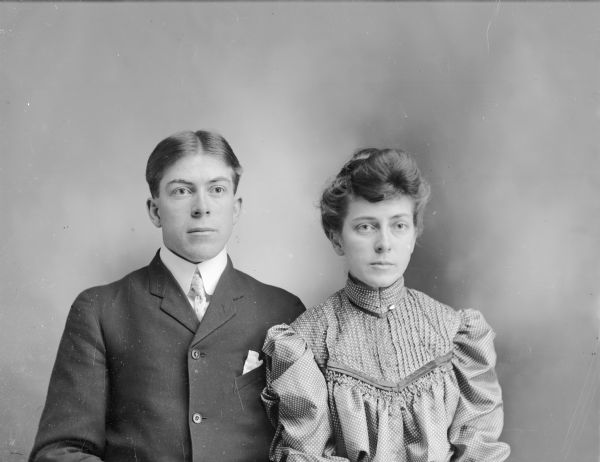Waist-up seated studio portrait of a young European American man and woman. The man is posed on the left wearing a dark-colored suit coat and light-colored necktie. The woman sits next to him on the right, wearing a ruffled blouse. Identified from left to right as Carl Ogden and Grace Ogden.