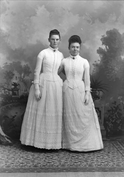Full-length studio portrait in front of a painted backdrop of two young European American women posed standing side-by-side with their arms around each other. They are both wearing gloves, light-colored dresses with lace trim, and collar pins. Identified from left to right as Bertine Benson and Amelia Torkelson.