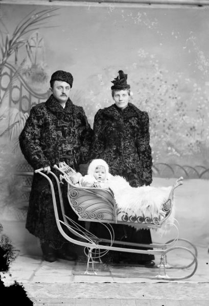 Studio group portrait in front of a painted backdrop of a European American family in winter costume and a baby carriage sleigh. The child is sitting in a sleigh covered by a light-colored fur and wearing a light-colored hooded coat. The man stands on the left wearing a dark-colored heavy winter coat and hat. The woman stands behind the sleigh on the right and is also wearing a dark-colored heavy winter coat, with a ribbon hat. Identified as probably Bill Maddox, his wife, and child.