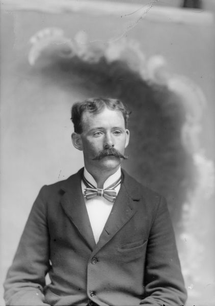 Waist-up studio portrait in front of a painted backdrop of a European American man with a moustache posed sitting and wearing a dark-colored suit coat and striped bow tie.
