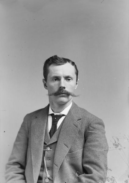 Waist-up studio portrait of a European American man with a moustache posed sitting. He is wearing a dark-colored suit coat, vest, watch fob, and necktie.