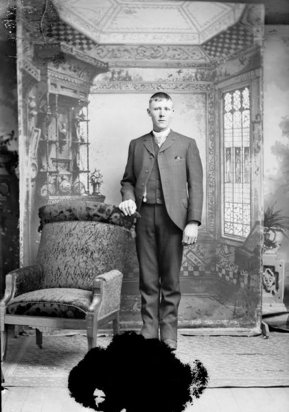 Full-length studio portrait in front of a painted backdrop of a young European American man posed standing with his right hand resting on the back of a chair. He is wearing a dark-colored suit coat, vest, ribbon watch fob, and light-colored necktie.