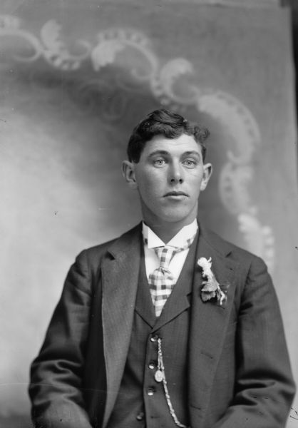 Waist-up studio portrait in front of a painted backdrop of a European American man posed sitting. He is wearing a dark-colored suit coat, vest, checkered necktie, watch fob, and floral boutonniere.