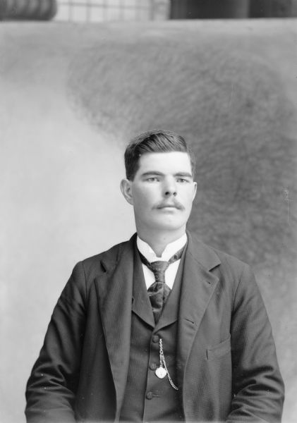 Waist-up studio portrait in front of a painted backdrop of a European American man with a moustache posed sitting. He is wearing a dark-colored suit coat, vest, paisley necktie, and watch chain.