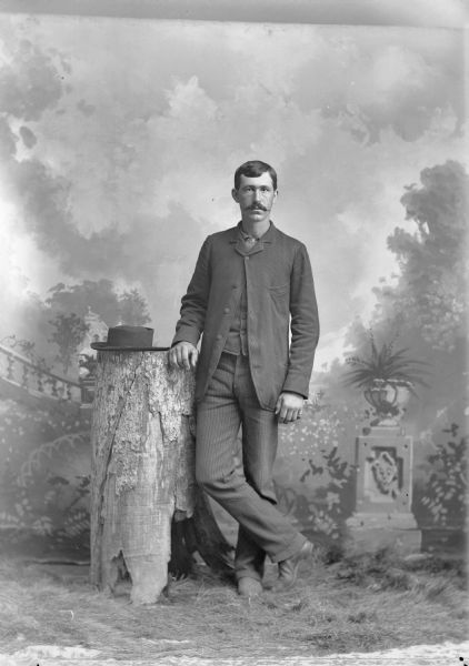 Full-length studio portrait in front of a painted backdrop of a European American man with a moustache. The man is posing standing with his right hand resting on a tree stump that a hat is resting on top of, and he is wearing a dark-colored suit coat, vest, and striped trousers.