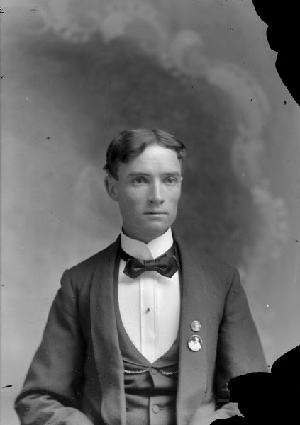 Waist-up studio portrait in front of a painted backdrop of a European American man posed sitting and wearing a dark-colored suit coat, vest, bow tie, watch chain. On his jacket lapel he is wearing two photograph buttons: one of an infant, the other of a woman.
