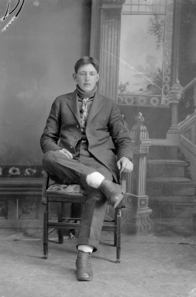 Full-length studio portrait in front of a painted backdrop of European American man posed sitting. He has his right ankle crossed over his left leg at the knee. He wears a dark-colored suit coat, turtleneck sweater, vest, and necktie.