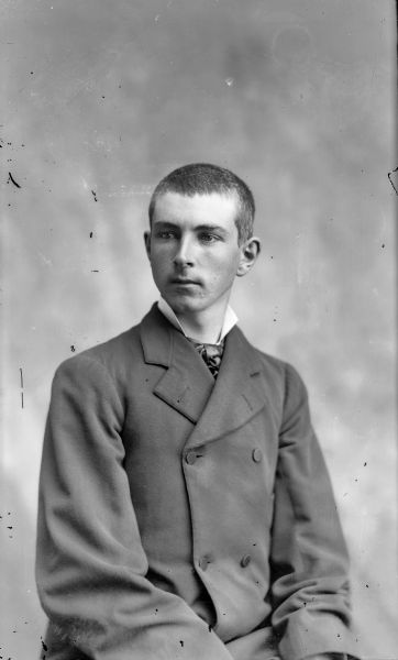 Waist-up studio portrait of a young European American man posed sitting. He is wearing a large dark-colored double-breasted suit coat and tie.