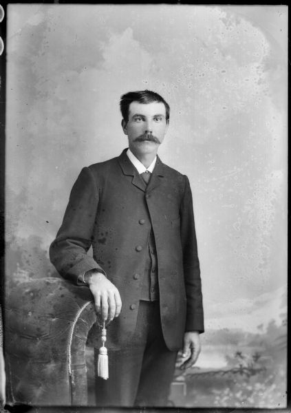 Studio portrait of a European American man with a moustache posed standing with his right arm resting on the back of a chair. He wears a dark-colored suit coat, necktie, and vest.