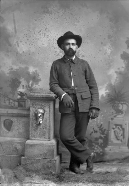 Full-length studio portrait in front of a painted backdrop of a European American man with a beard posed standing and leaning on a prop stone wall with his right arm. He wears a dark-colored belted coat, trousers, neckerchief, gloves and hat.