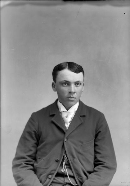 Waist-up studio portrait of a European American boy posed sitting. He is wearing a dark-colored suit coat, vest, light-colored necktie, tie pin, and watch chain.