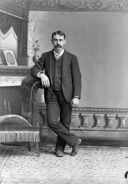 Full-length studio portrait in front of a painted backdrop of a European American man with a moustache. He is posed standing with his right arm resting on the back of a chair and his right leg crossed over his left leg. He is wearing a dark-colored suit coat, vest, trousers, watch chain, and long bow tie.
