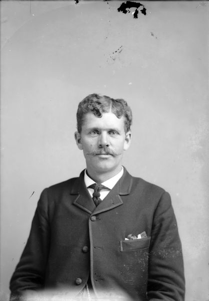 Waist-up studio portrait of a European American man with a moustache posed sitting. He is wearing a dark-colored suit coat, vest, watch chain, and necktie.