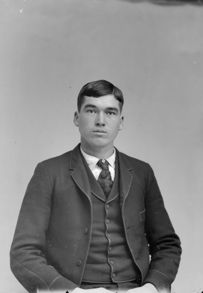 Waist-up studio portrait of a young European American man posed sitting. He is wearing a dark-colored suit coat, vest, necktie, and tie pin.
