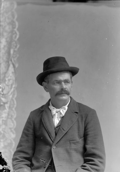 Waist-up studio portrait of a European American man with a moustache. He is posed sitting, and is wearing a dark-colored suit coat, vest, hat, light-colored shirt, and a long bow tie.