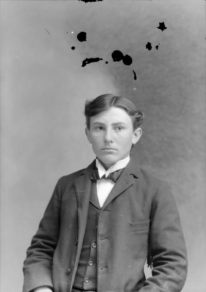 Waist-up studio portrait of a young European American man posed sitting. He is wearing a dark-colored suit coat, vest, and bow tie.