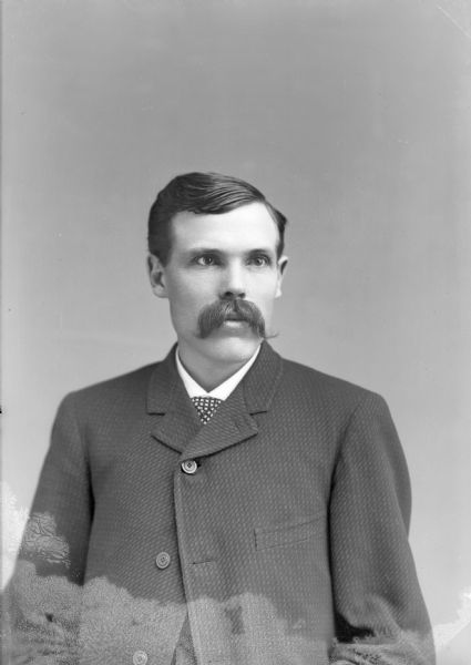 Waist-up studio portrait of a European American man with a moustache. He is posed sitting, and is wearing a dark-colored suit coat, vest, and necktie.