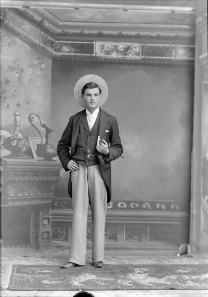 Full-length studio portrait in front of a painted backdrop of a young European American man posed standing and holding a walking stick under his left arm. He is wearing a dark-colored suit coat, light-colored trousers and neck tie, watch chain with a heart-shaped charm, and a straw hat.