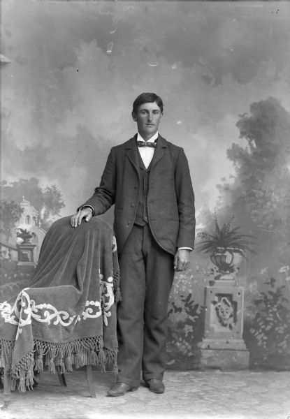 Full-length studio portrait in front of a painted backdrop of a European American man posed standing with his right hand resting on the back of a chair draped with a tasseled cloth. He is wearing a dark-colored suit coat, vest, and bow tie.