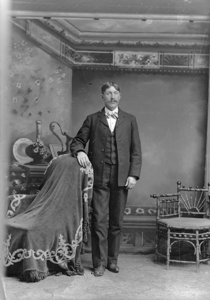 Full-length studio portrait in front of a painted backdrop of a European American with a moustache posed standing with his right arm resting on the back of a chair draped with a tasseled cloth. He is wearing a dark-colored suit coat, vest, striped light-colored bow tie, and watch chain. Another chair is on the right.