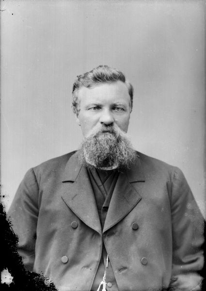 Waist-up studio portrait of a European American man with a beard posed sitting. He is wearing a dark-colored overcoat, suit coat, and watch chain.