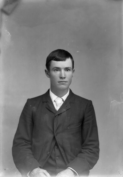Waist-up studio portrait of a young European American man posed sitting. He is wearing a dark-colored suit coat, vest, and light-colored necktie.