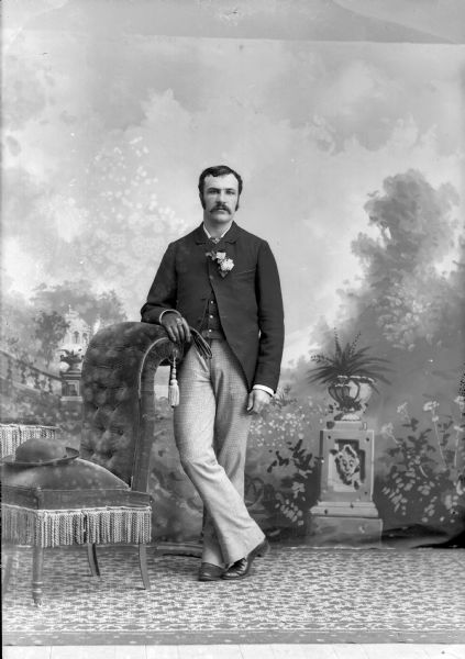 Full-length studio portrait in front of a painted backdrop of a European American man with a moustache posed standing with his right arm resting on the back of a chair. On the seat of the chair is his hat. The man is holding the left glove for his left hand in his right gloved hand. He is wearing a dark-colored suit coat, vest, tie, tie pin, and boutonniere.