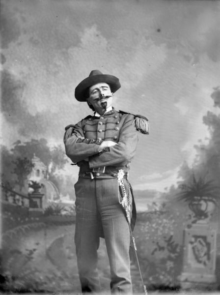 Three-quarter length studio portrait in front of a painted backdrop of a European American man with a moustache and goatee beard, possibly fake, posed standing with his arms crossed. He is wearing a military tunic with epaulettes, and a Civil War-era sword.