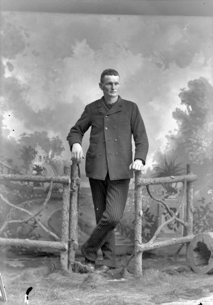 Full-length studio portrait in front of a painted backdrop of a European American man posed standing on fake grass in the opening of a prop wooden fence. He has both hands resting on the fence posts, and his left leg is crossed over his right leg. He wears a dark-colored suit coat, striped trousers, and a necktie.