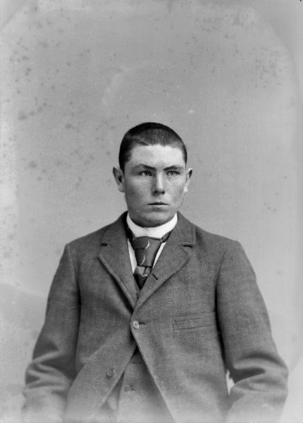 Waist-up studio portrait of a young European American man posed sitting. He is wearing a dark-colored suit coat, vest, and necktie.